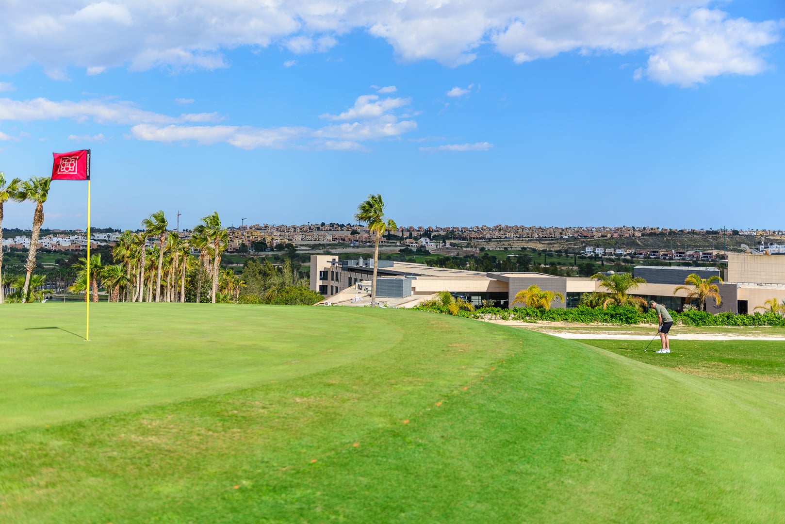 Sweeping view of a golf course green on a bright, partly cloudy day. Red flag with white logo on yellow flagpole on the left. Line of wind-blown palm trees behind flag. Golfer in light shirt and dark shorts on the right. Modern, light-colored clubhouse/resort building beyond the green. Sprawling residential area on distant hills under a blue sky with scattered clouds. Conveys openness, well-maintained course, and resort-style living.