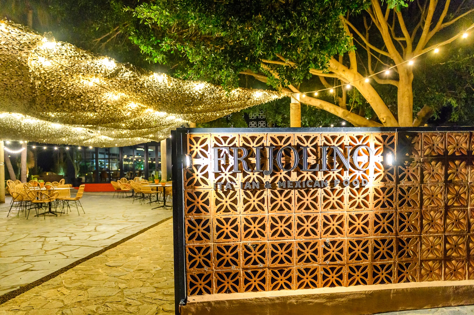 Entrance and outdoor seating of "FRIJOLINO ITALIAN & MEXICAN FOOD" restaurant at night. Metallic name on lit terracotta brick wall with floral/quatrefoil pattern. Flagstone patio with wicker chairs, tables. Camouflage netting canopy with warm string lights. Mature trees integrated into the setting. Creates a warm, inviting, rustic-chic ambiance for evening dining. Indoor restaurant section visible in background.