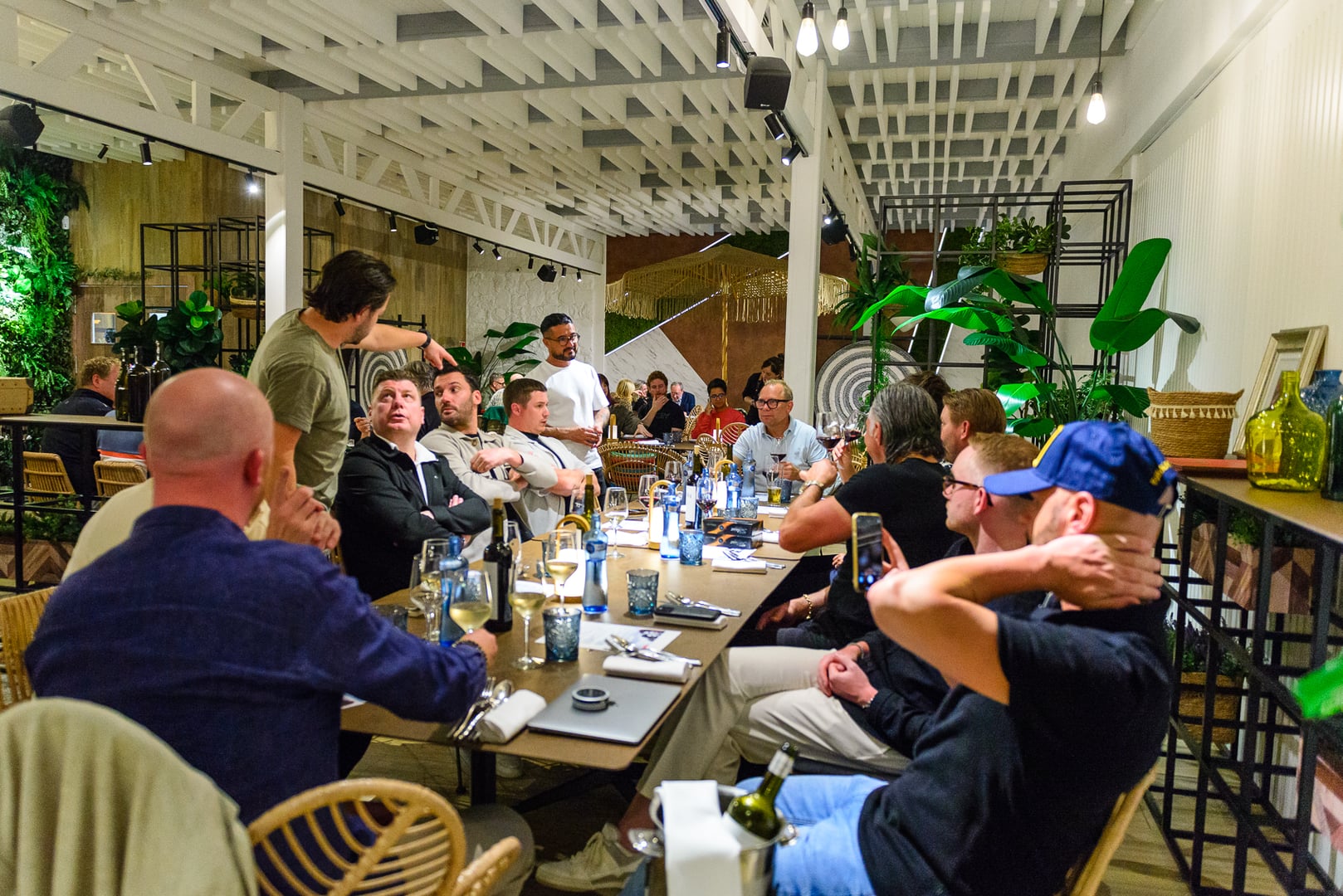 Lively dinner scene in a modern, well-lit restaurant. Group of mostly men in smart casual attire seated around a long table. Some conversing, others listening to one of two men standing. Table set with wine/water glasses, plates, cutlery, wine bottles, laptop. Contemporary decor: white coffered ceiling, track lighting, green wall, potted plants. Social and engaged atmosphere, suggesting a group or networking dinner.