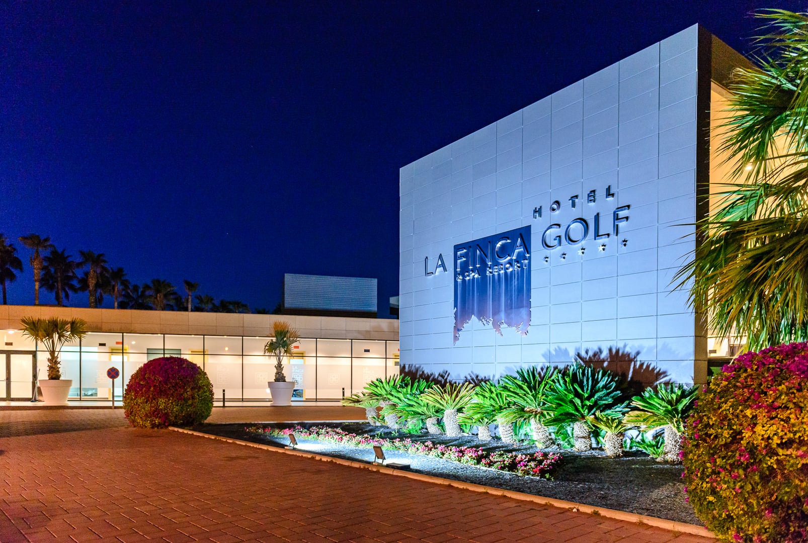 Exterior of "LA FINCA GOLF & SPA RESORT" hotel at night. Large, modern, illuminated pale-tiled facade with hotel name and logo. Deep indigo night sky. Foreground features red brick-paved area, landscaped with illuminated palm trees/cycads, and vibrant flowering bushes (bougainvillea). Illuminated glass windows and doors of another hotel section to the left. Evokes modern luxury and a welcoming resort ambiance at night.