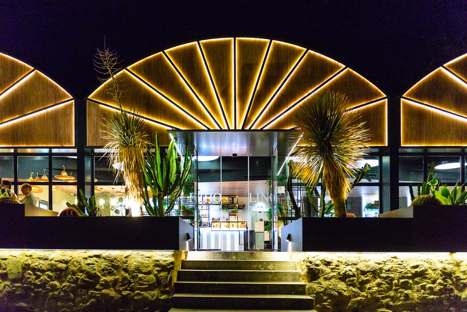 Striking night view of Frijolino restaurant entrance. Facade features large, backlit semi-circular wooden panel (sunburst effect) and similar flanking panels. Glass doors reveal lit interior bar/reception with "FRIJOLINO ITALIAN & MEXICAN FOOD" visible. Raised terrace with desert plants (cacti, yuccas) in dark planters. Concrete stairs lead up from a rugged, illuminated stone wall base. Stylish, contemporary, and inviting ambiance with sophisticated lighting.