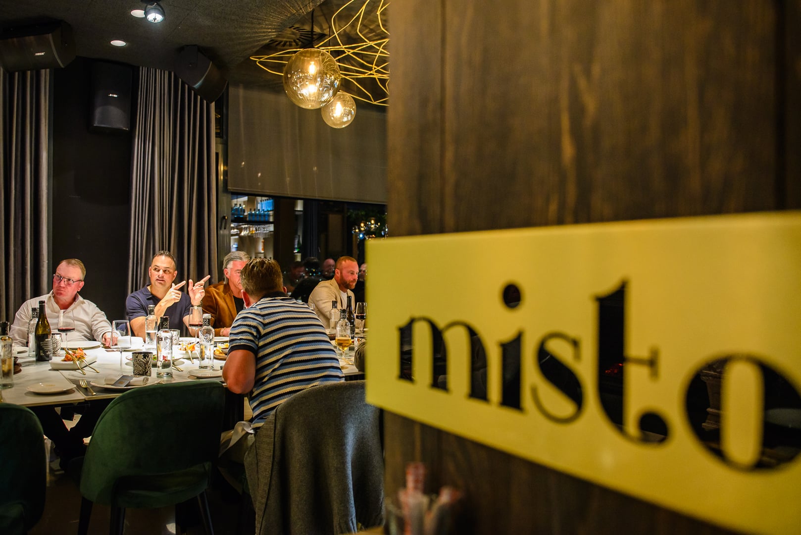 View of "misto" restaurant interior. Prominent yellow, backlit sign with stylized cutout letters "misto" on dark wooden pillar in foreground. Background shows group of mostly men at a dining table, conversing. Sophisticated ambiance, dark walls, grey curtains, modern spherical pendant lights with gold wire design. Table set for dinner. Upscale, intimate dining experience.