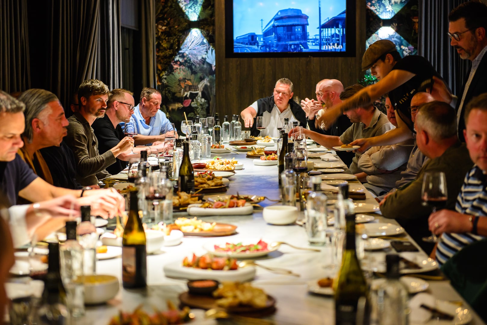 Large group of men at a long, white-clothed dining table in Misto restaurant. Table filled with small plates of food (tapas/shared meal), wine/water bottles. Men in smart casual attire, eating and conversing. Waiter serving a guest. Decor: dark walls, grey curtains, botanical/tropical print wallpaper. TV screen shows "misto" logo and old train. Lively, convivial atmosphere of an upscale social gathering.