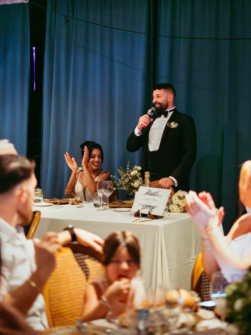 De bruidegom houdt een speech aan de eretafel terwijl de bruid applaudisseert.